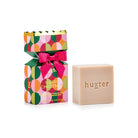 Huxter bon bon box patterned with green, pink and orange circles, pink bow and gold scalloped top, beside the pale cleansing bar stamped huxter.