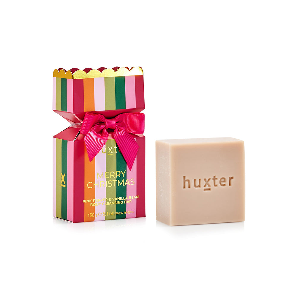 Huxter bon bon box with pink, green, orange and red stripes, pink bow and gold scalloped top, beside the pale cleansing bar stamped huxter.