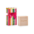 Huxter bon bon box with pink, green, orange and red stripes, pink bow and gold scalloped top, beside the pale cleansing bar stamped huxter.