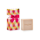 Huxter bon bon box with pink, orange and green diamonds, pink bow and gold scalloped top, beside the pale cleansing bar stamped huxter.