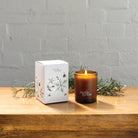 The Fir, Spruce & Pine candle box and the lit amber glass candle on a wooden table with pine sprigs.