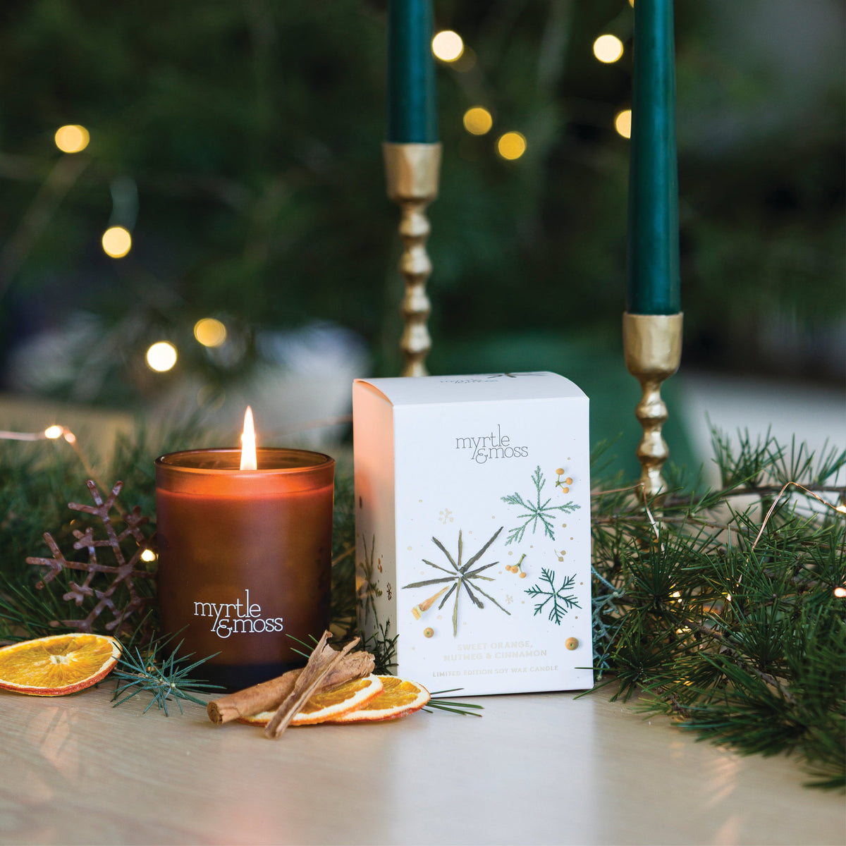 Lit Myrtle & Moss Sweet Orange, Nutmeg & Cinnamon candle in an amber glass jar beside its white box printed with green and gold snowflakes, among pine branches.