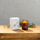 The Sweet Orange, Nutmeg & Cinnamon candle box and the lit amber glass candle on a wooden table with pine sprigs.