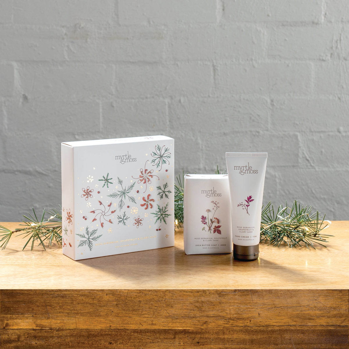The Rose Geranium soap and hand cream gift box beside the soap bar and hand cream tube on a wooden table.