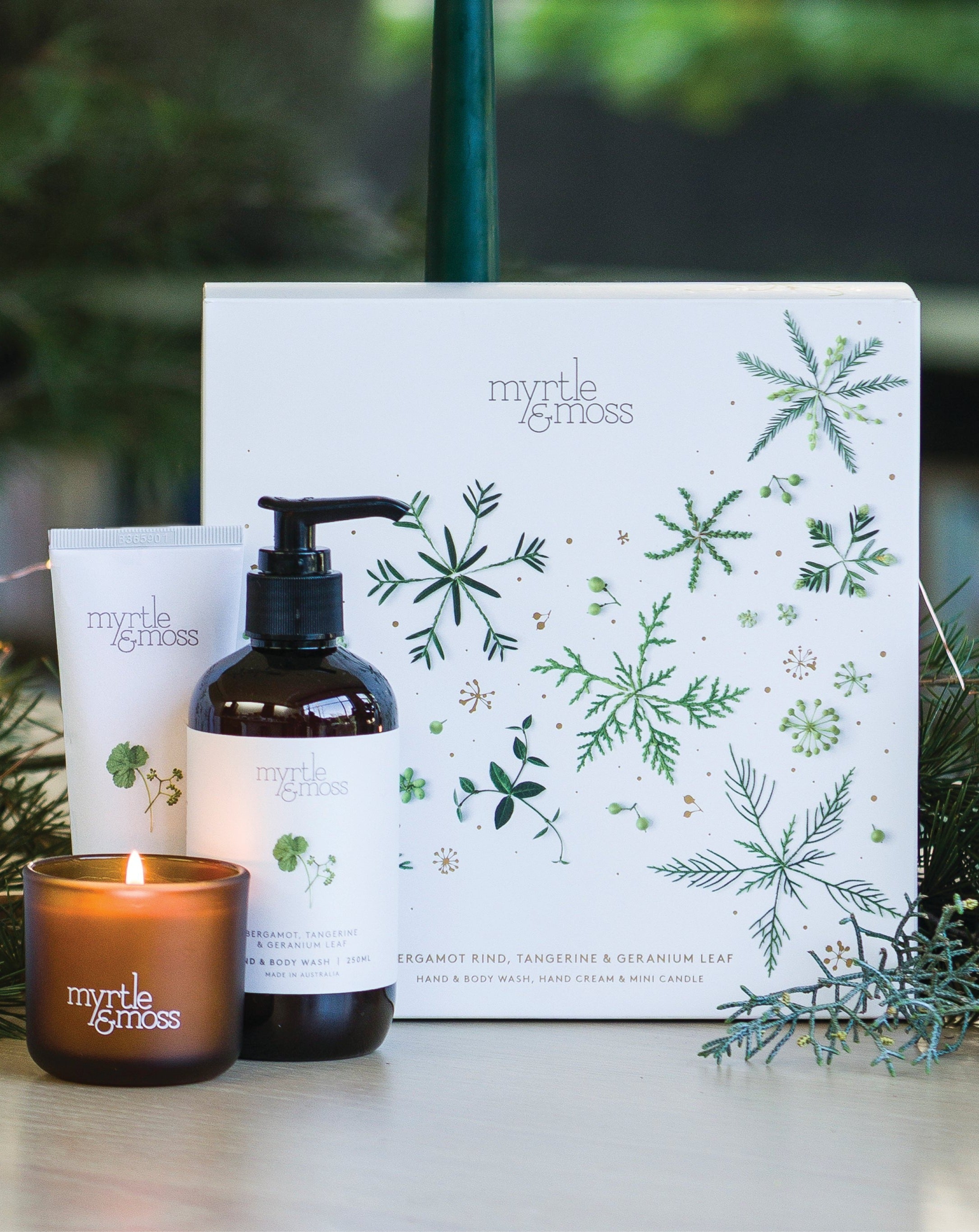 The Bergamot trio's lit mini candle, hand cream and hand and body wash in front of the gift box, with pine branches.
