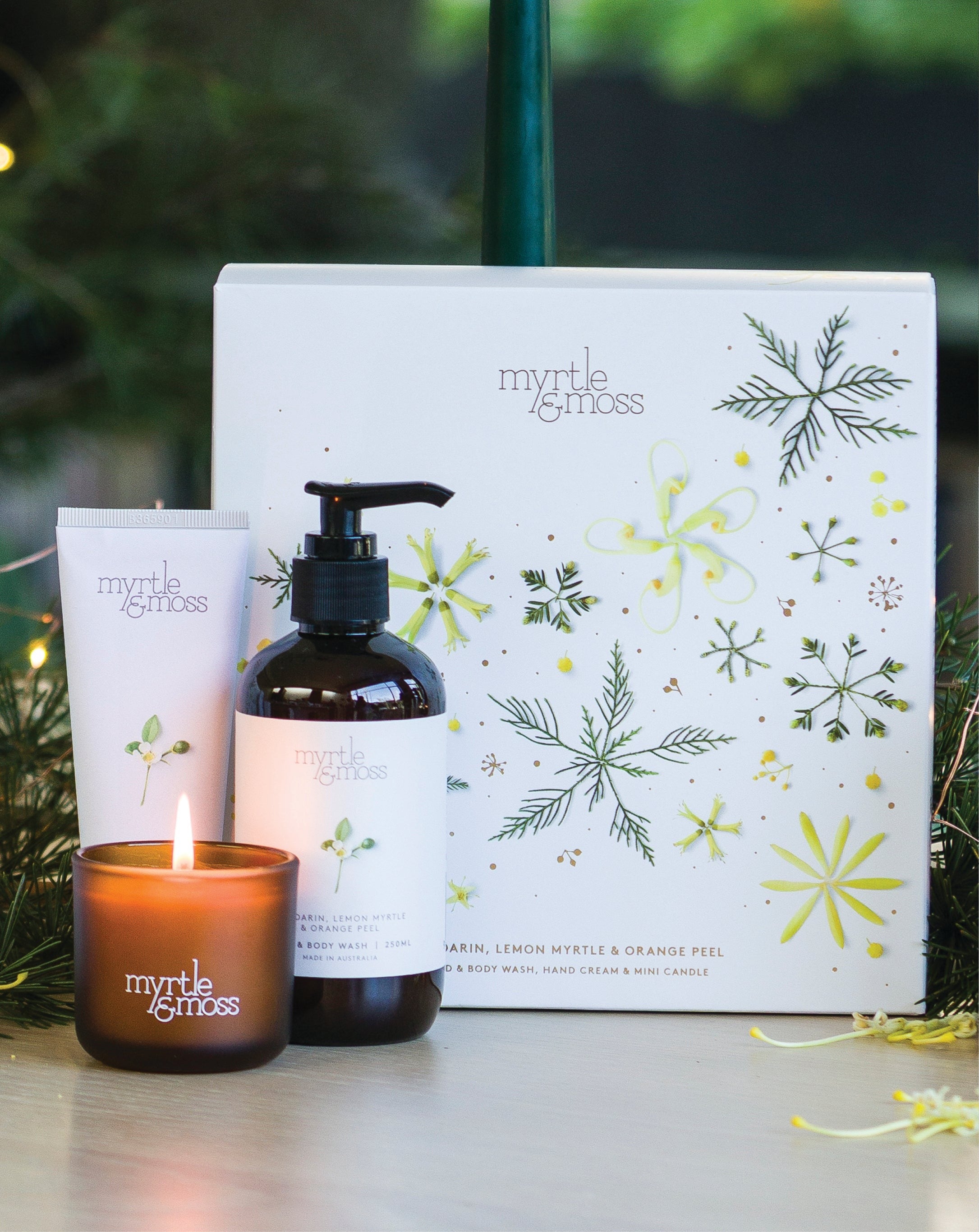 The Citrus trio's lit mini candle, hand cream and hand and body wash in front of the gift box, with pine branches.