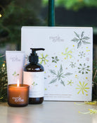 The Citrus trio's lit mini candle, hand cream and hand and body wash in front of the gift box, with pine branches.