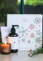 The Rose Geranium trio's lit mini candle, hand cream and hand and body wash in front of the gift box, with pine branches.
