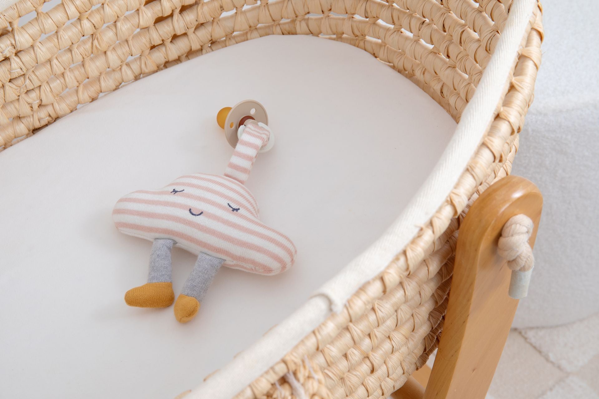 The cloud pacifier holder lying in a woven bassinet with a dummy on a wooden ring attached to its tab.