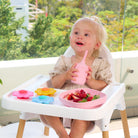 Toddler in a high chair with a pink suction plate and daisy spinners on the tray