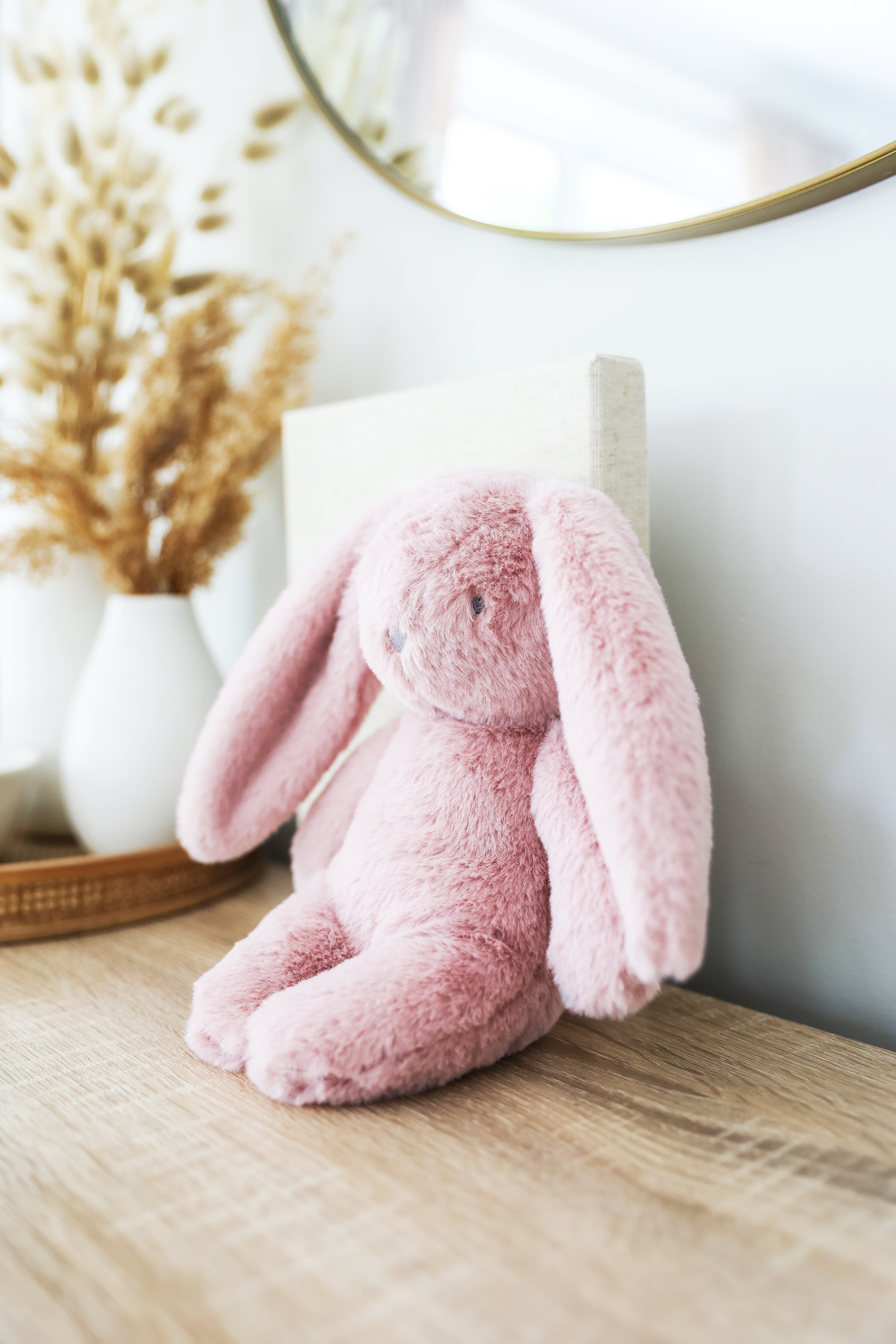 Pink plush bunny sitting on a timber shelf beside a vase of dried grasses
