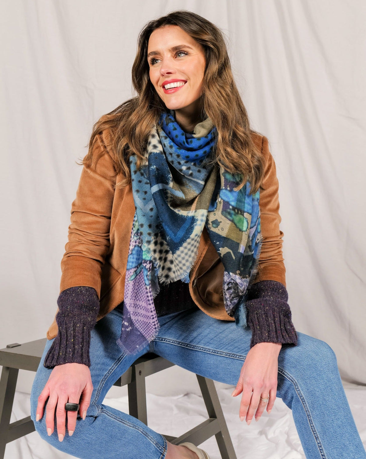 Smiling model seated in jeans with the blue print scarf wrapped at her neck