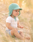 Baby sitting in long grass wearing the green striped Mini Pistachio flap cap