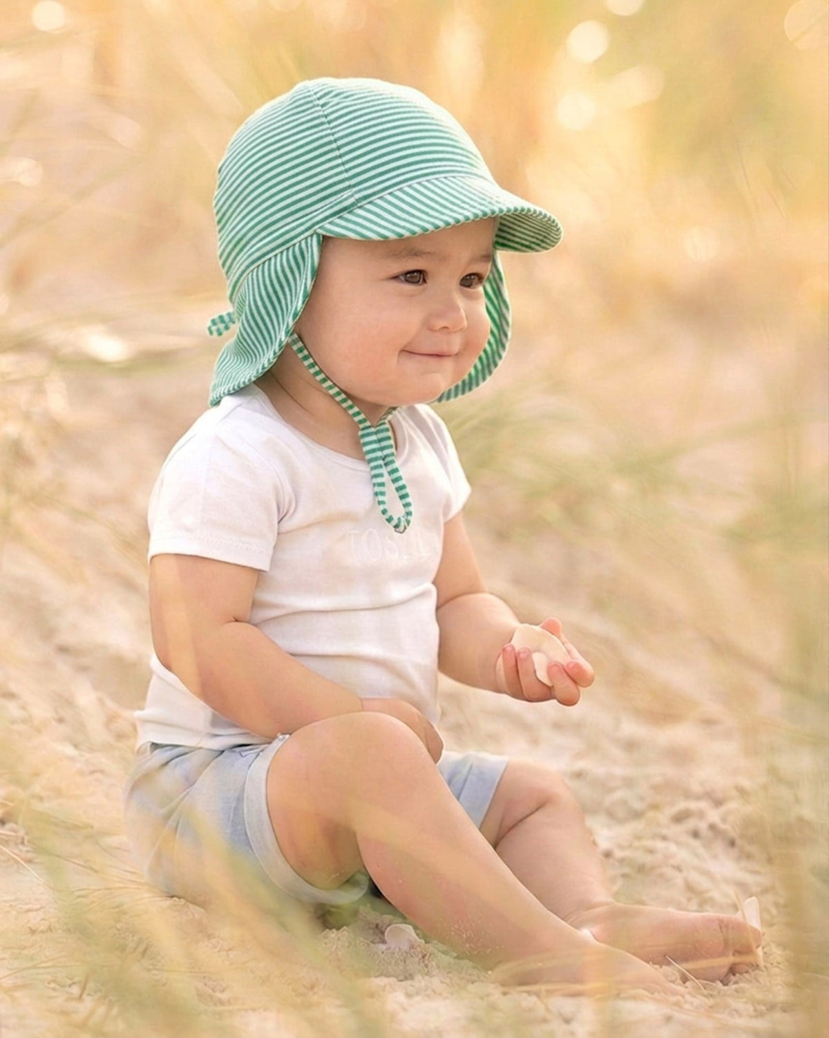 Baby sitting in long grass wearing the green striped Mini Pistachio flap cap