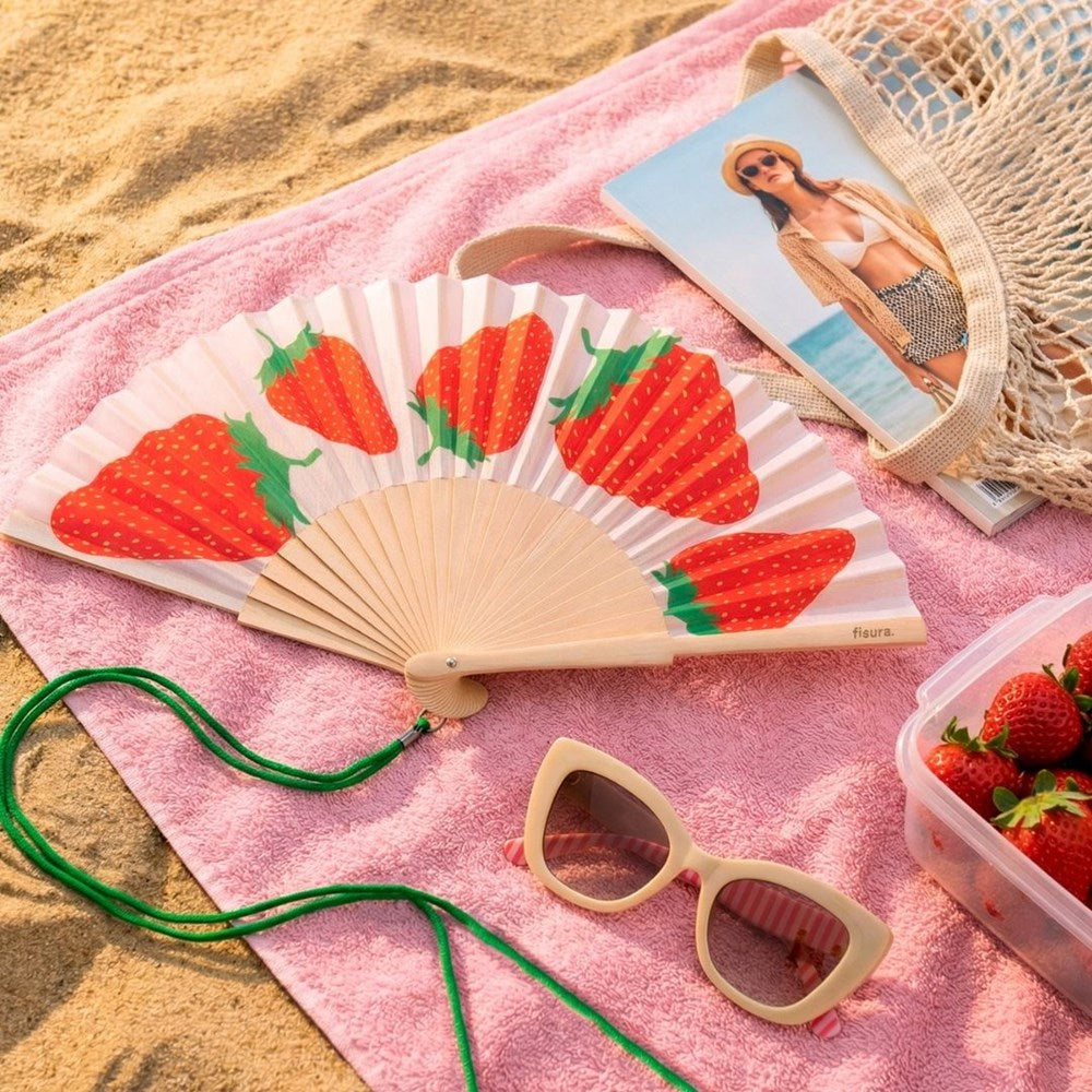 Strawberry fan lying open on a pink towel beside sunglasses and a magazine