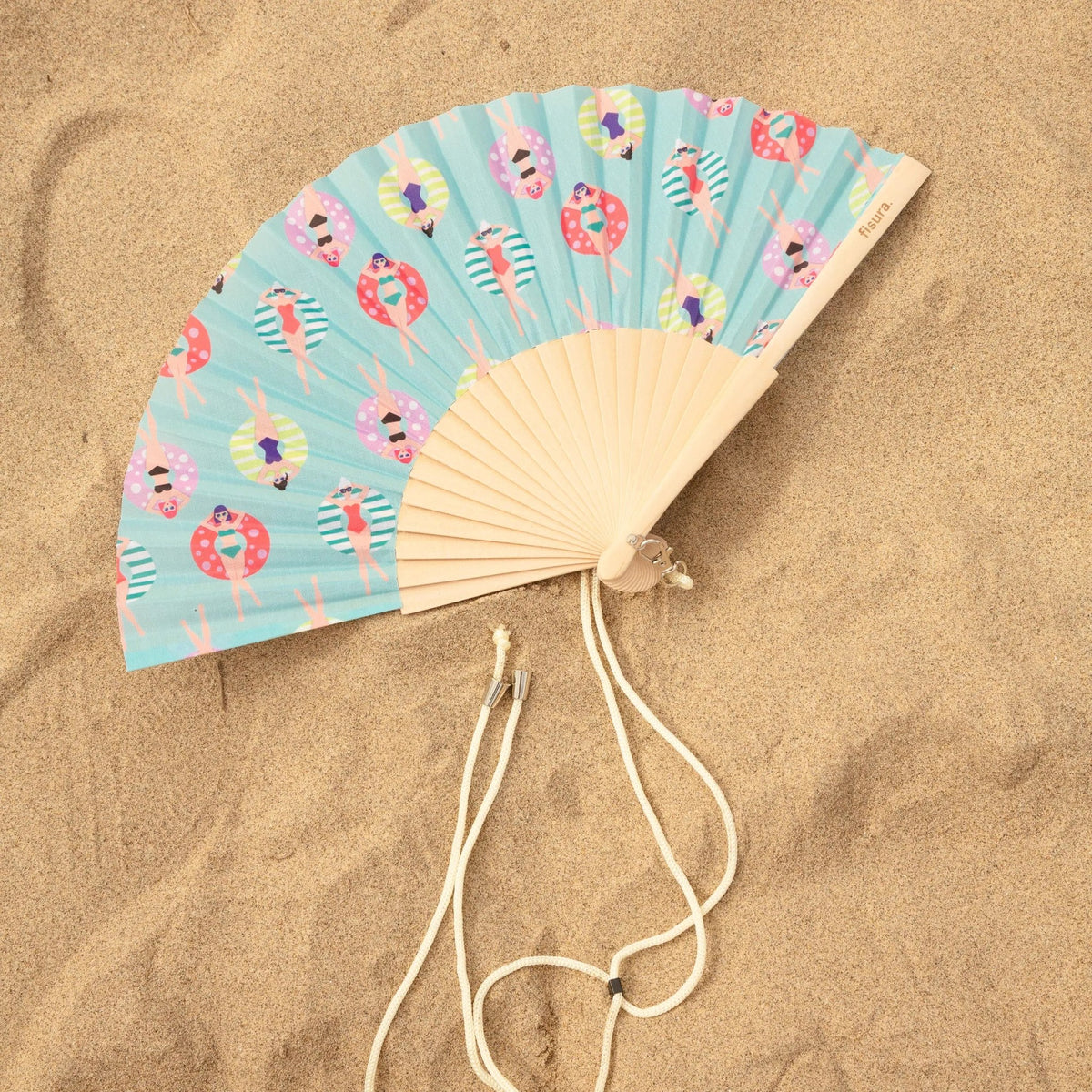 The Swimmers fan open on sand: small swimmers printed on aqua, with its timber ribs and cord.