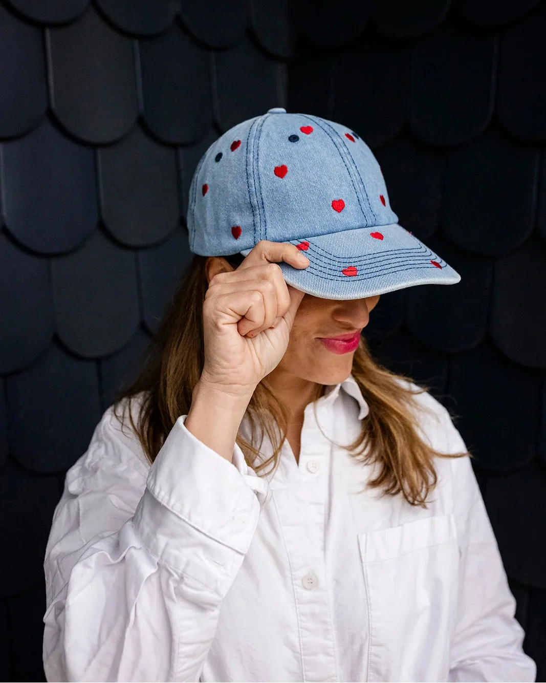 Woman in a white shirt tilting the red-heart denim cap down over her eyes against a dark shingled wall.