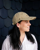 Woman in a white shirt wearing the khaki dachshund cap in profile against a dark shingled wall.