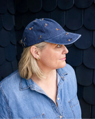 Woman in a denim shirt wearing the navy dachshund cap in profile against a dark shingled wall.
