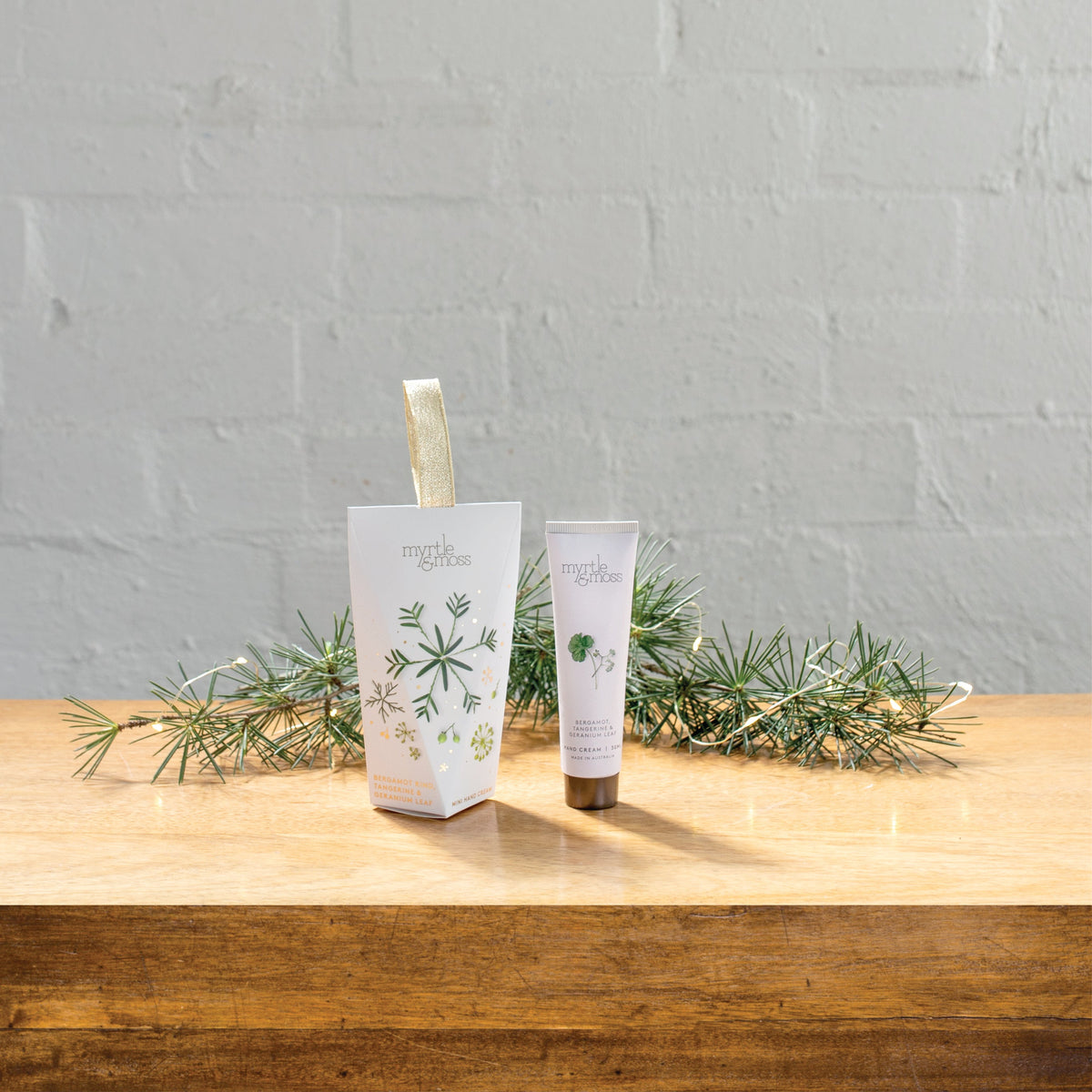 The Bergamot hanging hand cream box beside its 30 mL tube on a wooden table with pine sprigs.