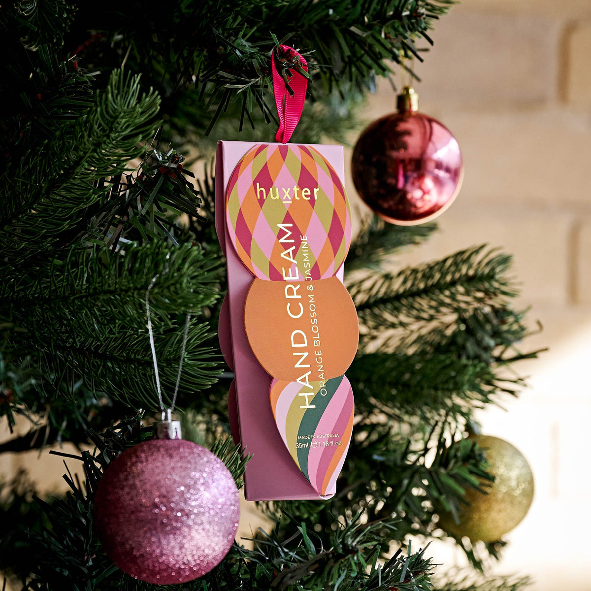 The pink Huxter hand cream box hanging by its ribbon on a Christmas tree beside pink and gold baubles.