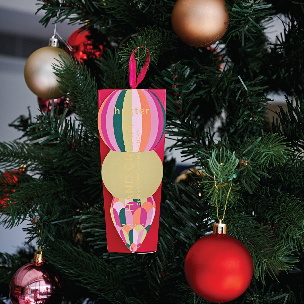 The red Huxter hand cream box hanging by its ribbon on a Christmas tree among red and gold baubles.