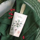 Myrtle & Moss Rose Geranium hanging hand cream box with a gold ribbon loop, printed with a red and green snowflake and red berries, on a green napkin.