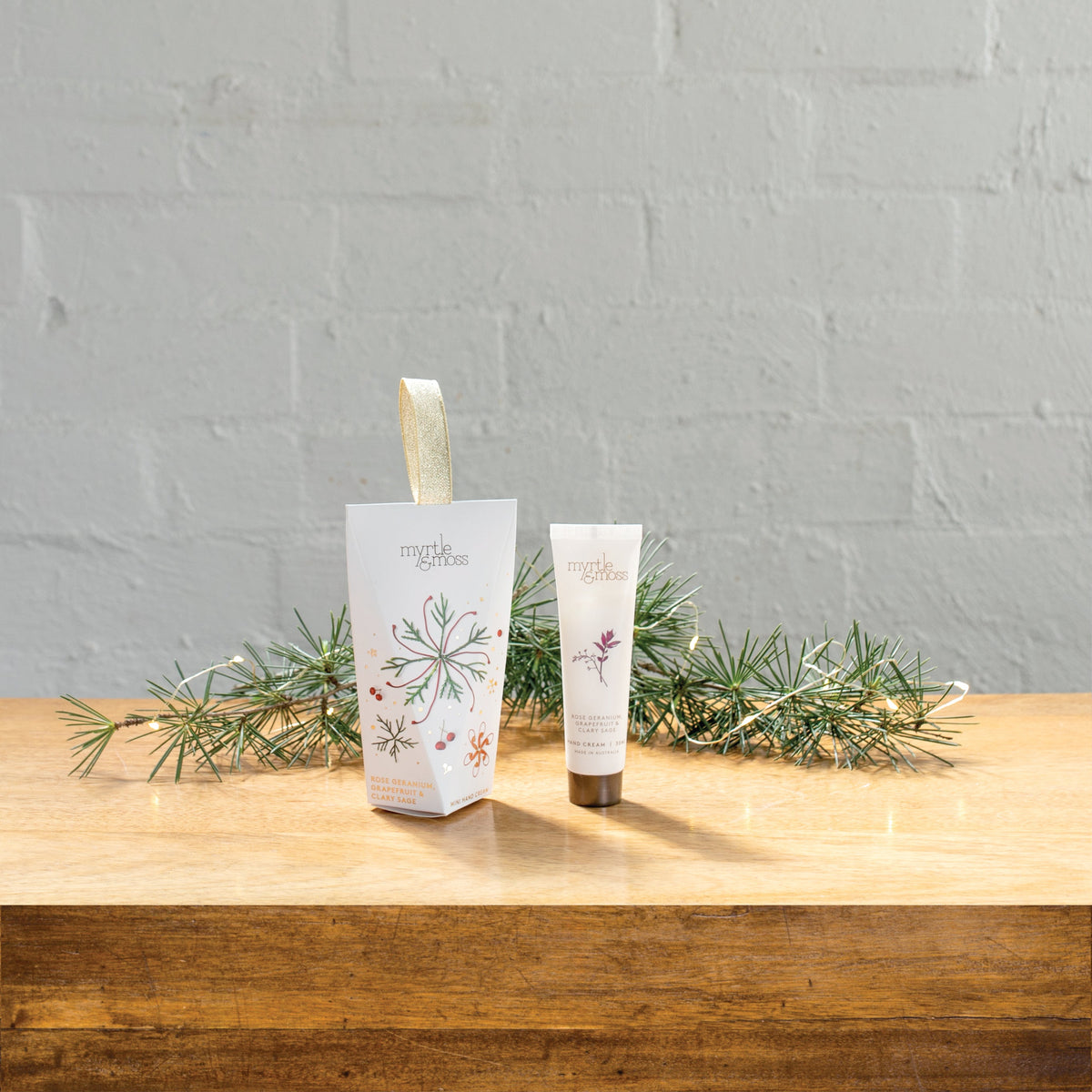 The Rose Geranium hanging hand cream box beside its 30 mL tube on a wooden table with pine sprigs.