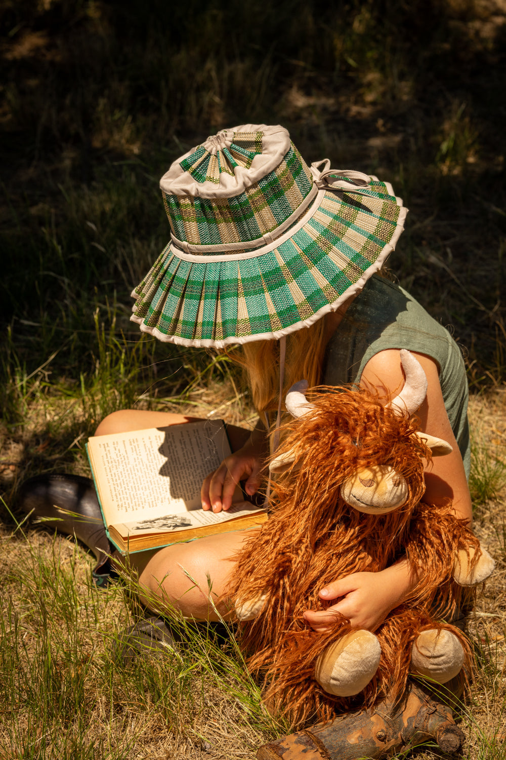 A child in a checked bucket hat reading in long grass with the cow on their lap.