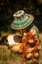 A child in a checked bucket hat reading in long grass with the cow on their lap.