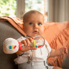 Baby held against an orange cushion, gripping the maracas rattle in both hands.