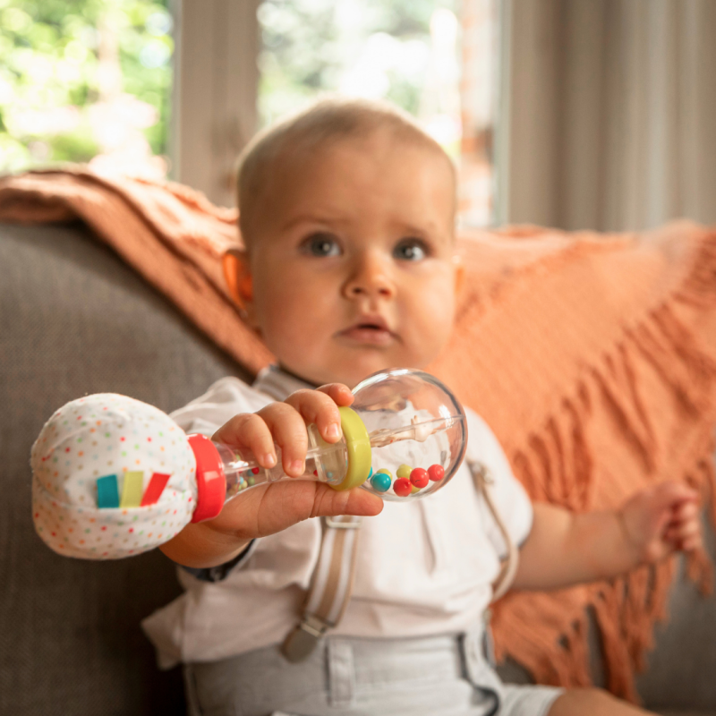 Baby held against an orange cushion, gripping the maracas rattle in both hands.