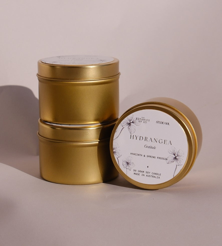 Three gold Hydrangea travel tins, one tipped forward to show its lid label with a drawing of the hydrangea.