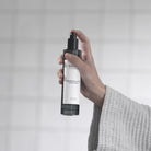 A hand spraying the smoked glass Lemongrass & Lime Room Spray in front of a tiled wall.