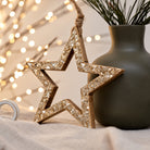 Open timber star with a gold flaked face and a jute hanging loop, leaning against a sage green vase in front of fairy lights