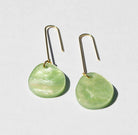 A pair of long gold hook earrings, each hung with a flat round disc of soft green marbled acrylic.