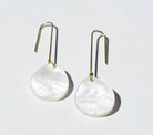 A pair of long gold hook earrings, each hung with a flat round disc of white pearlescent marbled acrylic.