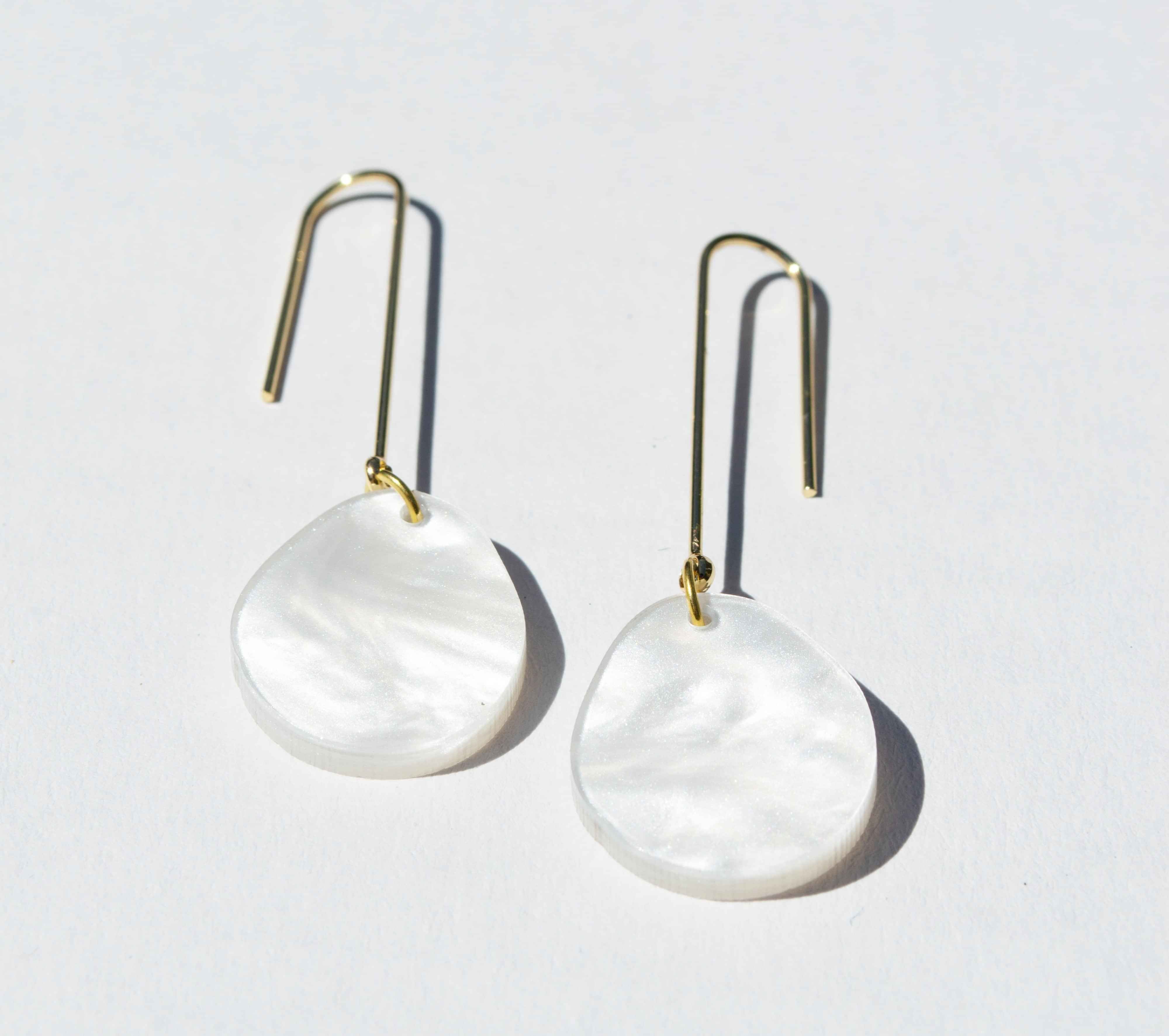 A pair of long gold hook earrings, each hung with a flat round disc of white pearlescent marbled acrylic.