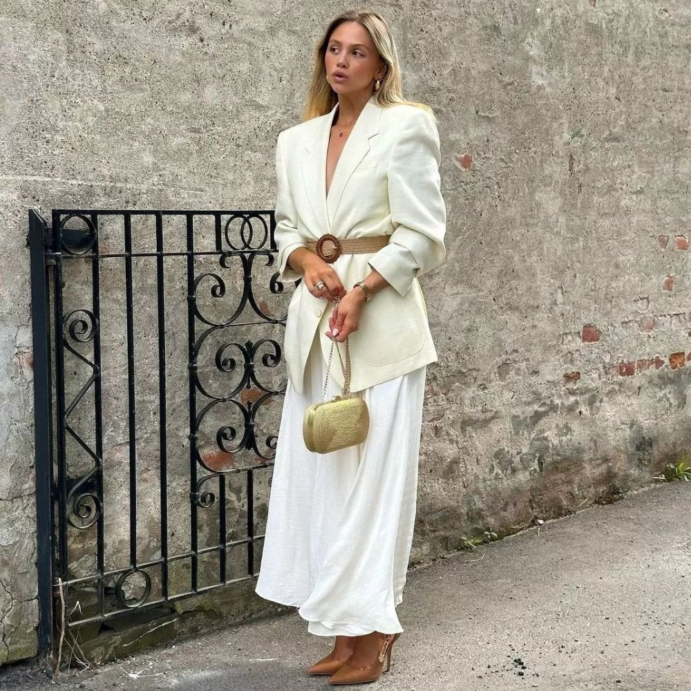 Woman in a cream blazer and long white skirt beside a wrought-iron gate, the gold clutch hanging from her hand