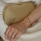 Hand wearing a gold bangle and ring resting on the gold clutch against cream satin