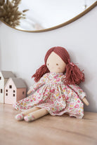 Matilda sitting against a wall in her pink floral dress, beside two little timber houses.