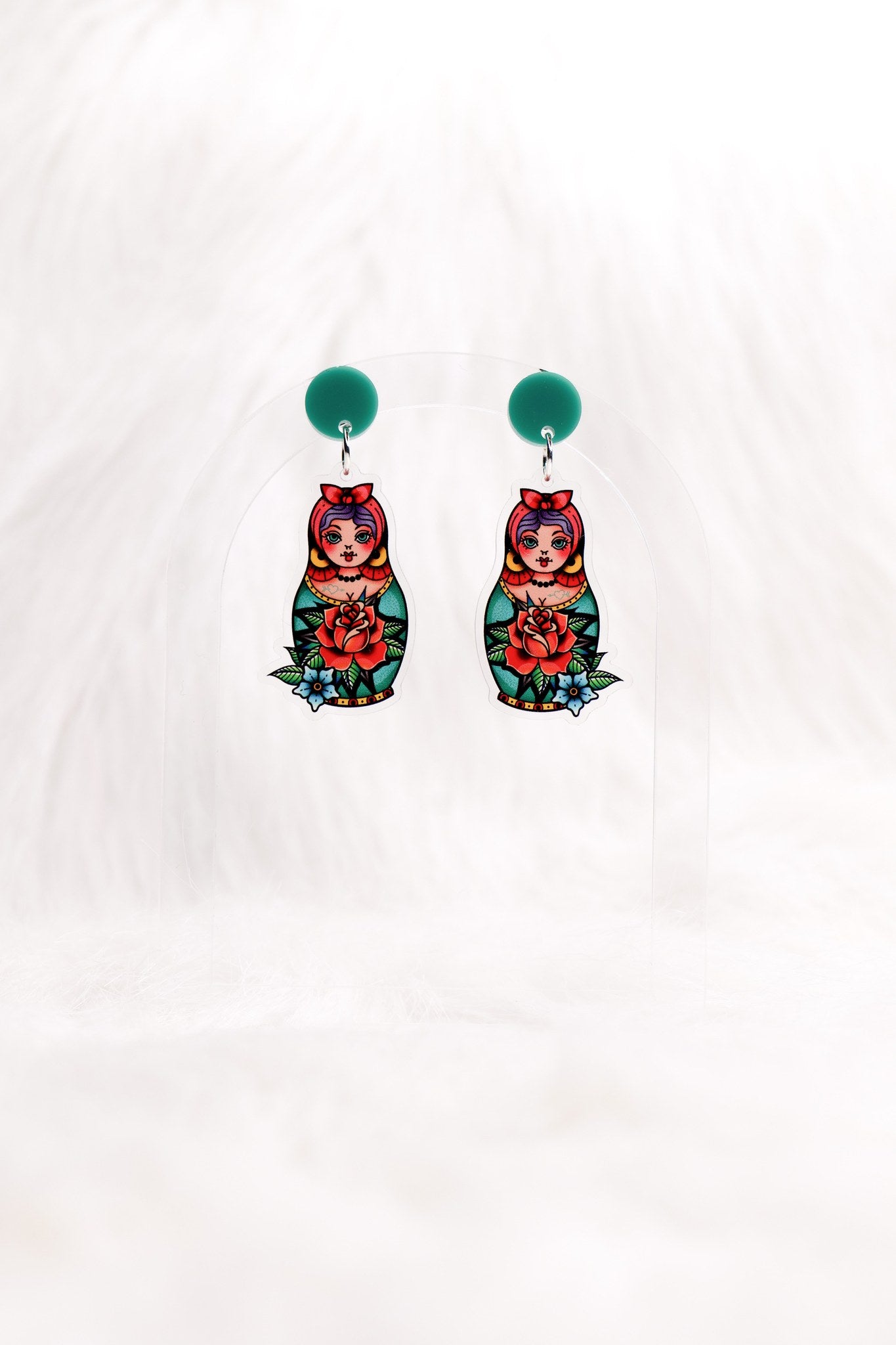 The Matryoshka earrings hanging on a clear stand: green studs above painted nesting dolls.