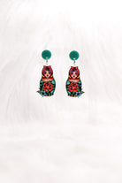 The Matryoshka earrings hanging on a clear stand: green studs above painted nesting dolls.