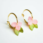 A pair of gold hoop earrings, each hung with a small soft pink flower and a slender green leaf in marbled acrylic.