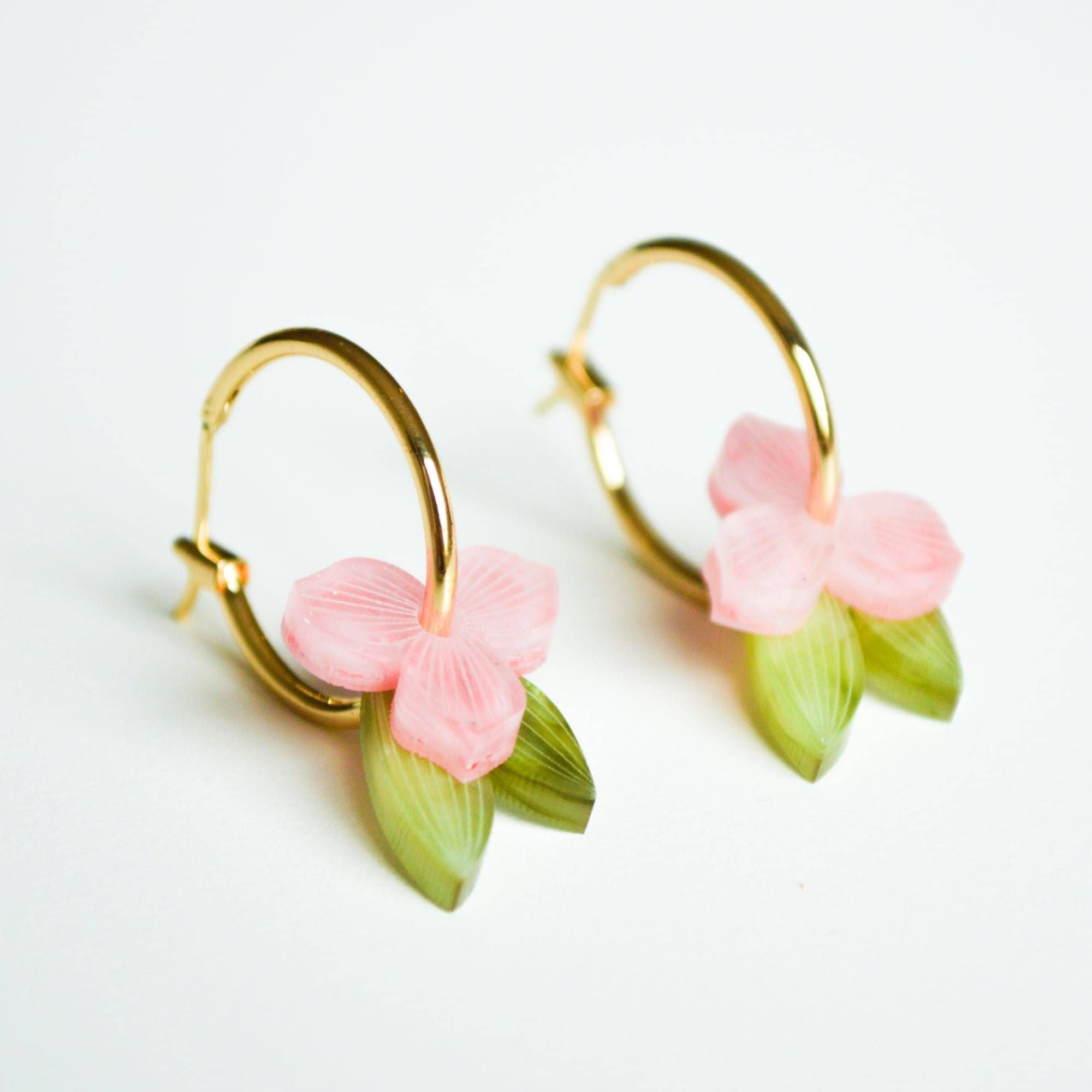 A pair of gold hoop earrings, each hung with a small soft pink flower and a slender green leaf in marbled acrylic.