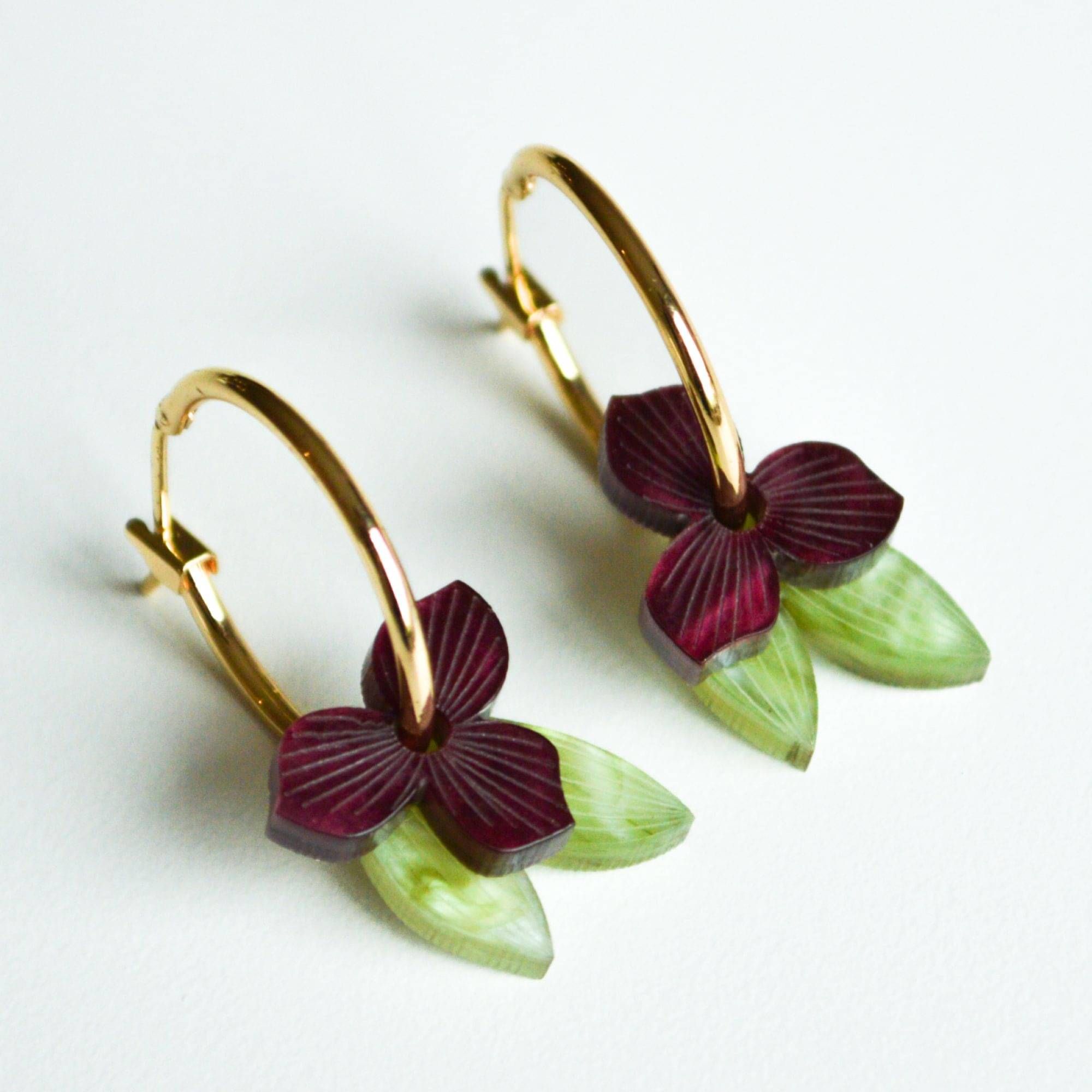 A pair of gold hoop earrings, each hung with a small deep burgundy flower and a slender green leaf in marbled acrylic.