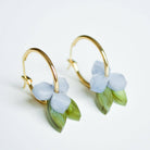 A pair of gold hoop earrings, each hung with a small pale periwinkle blue flower and a slender green leaf in marbled acrylic.