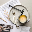 Black and cream wick trimmers beside a lit candle on a speckled ceramic plate