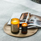 Two lit Mojo candles on a round wooden board over an open magazine on grey herringbone