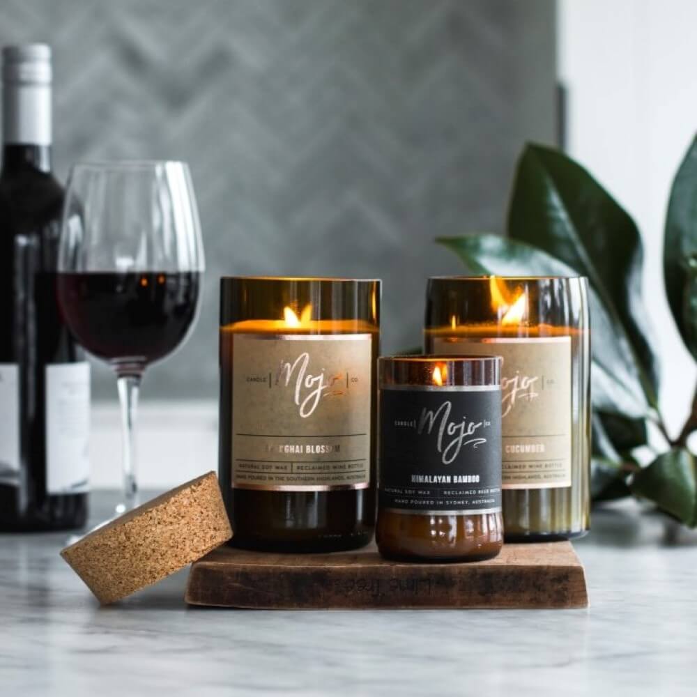 Three lit Mojo candles of different sizes on marble with a glass of red wine and green foliage